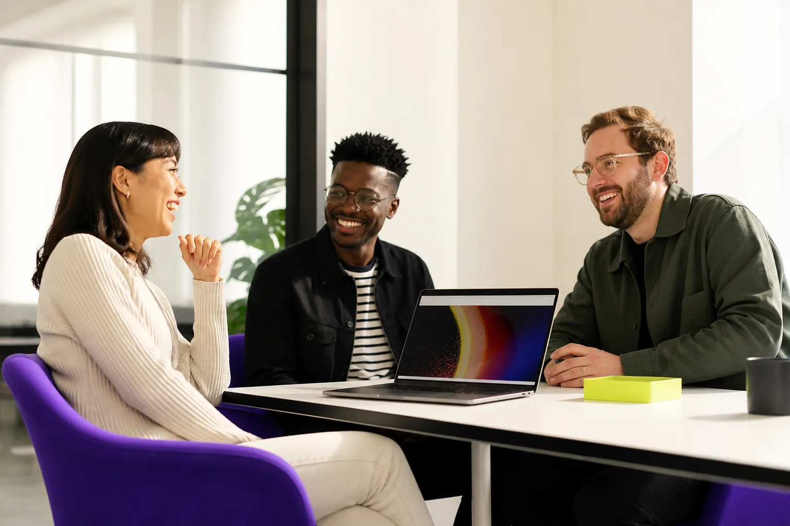 Three colleagues smiling together around a table in a bright creative office.